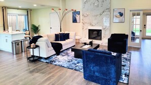 Una sala de estar moderna con un sofá blanco, una mesa de centro negra, un sillón de terciopelo azul, una chimenea y un ventilador de techo.