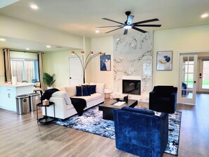 A modern living room with a white sofa, a black coffee table, a blue velvet chair, a fireplace, and a ceiling fan. - 3-BDR Resort-style Home - Hot Tub, Mini Golf, Fire Pit, Grill, 1Gig WiFi, Office (Richardson)