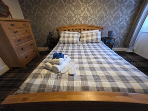 A neatly made bed with a checkered bedspread, two pillows, a wooden dresser with drawers, and a nightstand with a lamp.