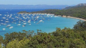 A coastal area with numerous boats anchored in clear blue waters, surrounded by dense green vegetation and distant mountains. - Family Escape Steps From the Sea (Hyères)