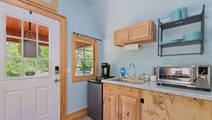 Cocina compacta con puerta blanca, armarios de madera, horno integrado, microondas y ventana con vistas al verdor.