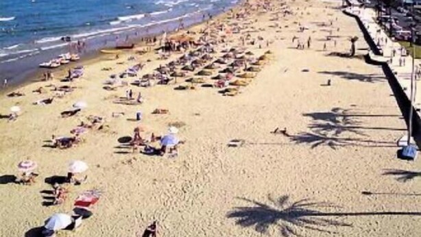 Uma praia com pessoas tomando sol, um afloramento rochoso na água e edifícios ao longo da costa.