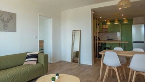 A modern living room with a sofa, a dining table with chairs, a mirror, and a kitchen area with green cabinets and yellow pendant lights. - Private Room in a Shared Apartment - Bron (Bron)