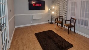A living room with a flat-screen TV mounted on the wall, a dark rug, wooden flooring, two wooden chairs, and a patterned curtain. - Designer 5-Bedroom Home by Elizabeth Line (Greater London)
