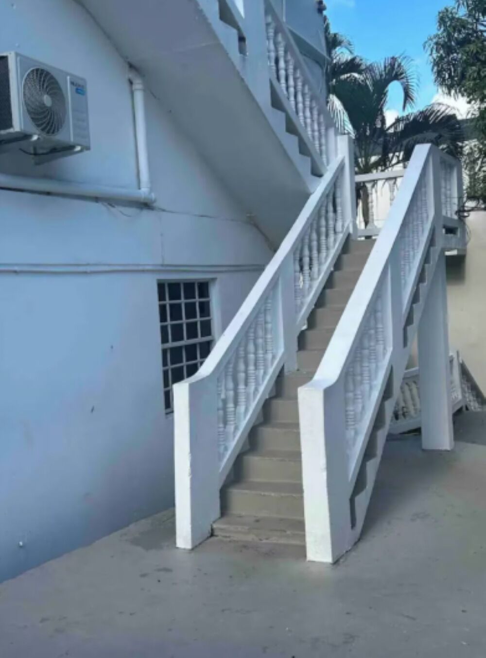 Una escalera conduce a un edificio de fachada blanca, con barandilla metálica y un equipo de aire acondicionado en la pared.