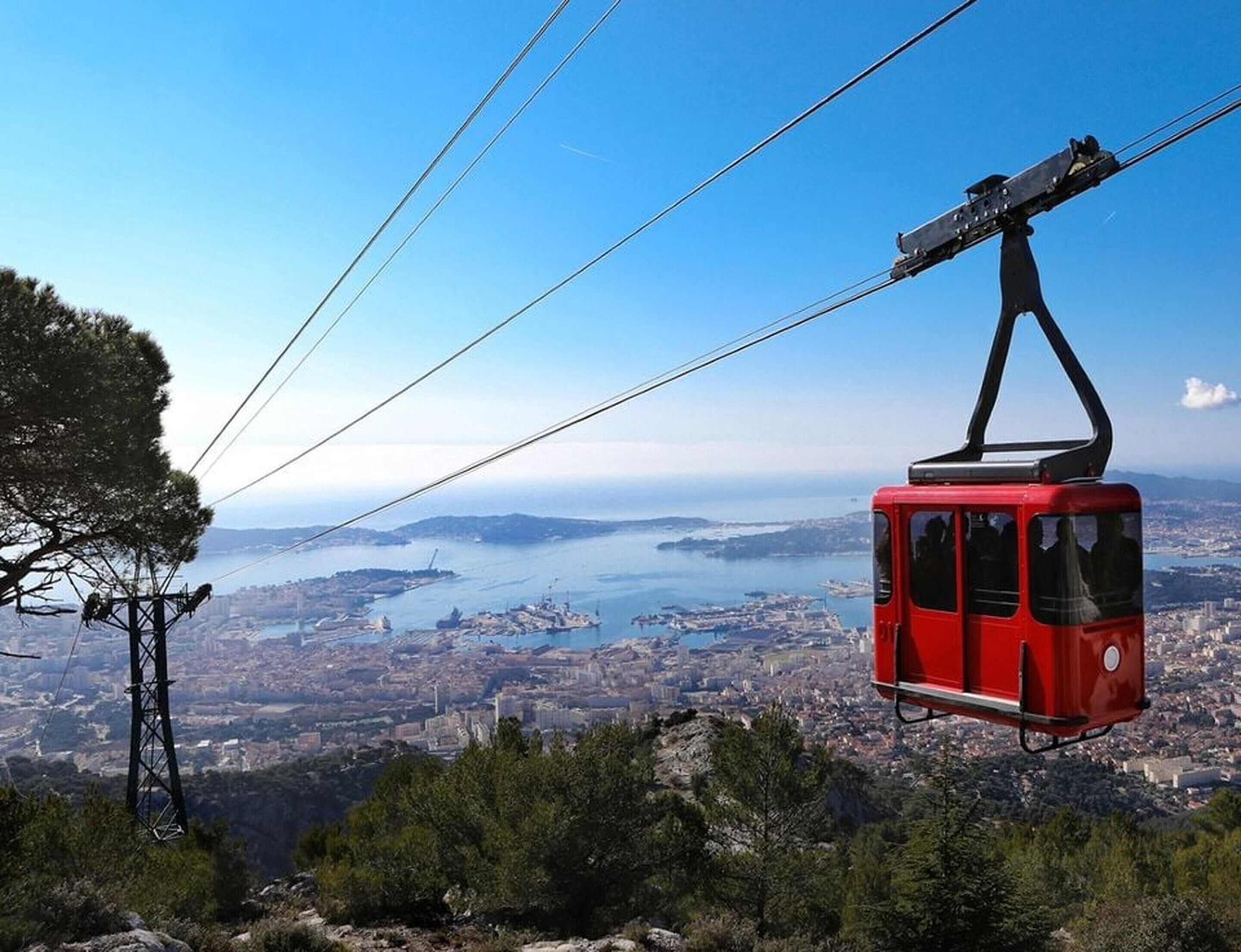 A red cable car suspended over a city with a view of the coastline and hills.
