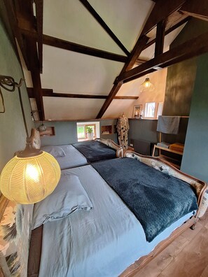 Une pièce avec un plafond en pente, des poutres en bois, deux lits avec une literie aux couleurs sombres et claires, un luminaire suspendu et une petite fenêtre.