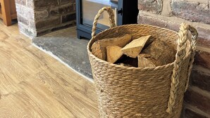 Een kamer met een open haard van baksteen, een gevlochten mand met houtblokken, en een houten vloer.