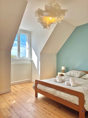 Ein Schlafzimmer mit schräg versiegelter Decke, großem Fenster, Heizkörper, einem hölzernen Bett und einem Nachttisch mit Lampe.