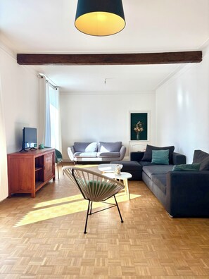 Ein Wohnzimmer mit Holzboden, einer Couch, einem Sessel, einem kleinen Tisch, einem Fernseher auf einer Holzkommode und einer Pendelleuchte.