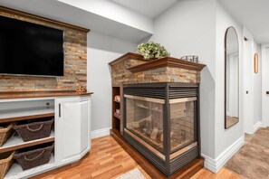 A living room with a stone fireplace, a television mounted above it, and a white cabinet with baskets. - 1 BR Executive Suite | Laundry, Parking & Workspace (Smiths Falls)