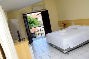 Habitación de hotel con cama, escritorio, silla, una pequeña mesa con un jarrón de flores y un balcón con vistas a zonas verdes y edificios.
