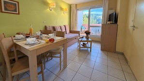 Ein Essbereich mit einem Tisch für zwei Personen, einer Couch mit Kissen, einem kleinen Tisch mit einer Vase und Blick durch ein Fenster.