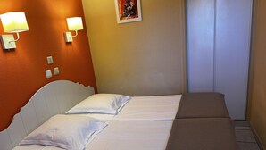 Un letto rifatto con lenzuola bianche e una coperta marrone, due lampade a parete, un piccolo quadro e una porta bianca.