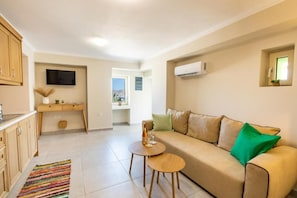 A living room with a sofa, a small table, a TV mounted on the wall, and a kitchen area with cabinets and a window. - Sunrise Apartments (Karpathos)