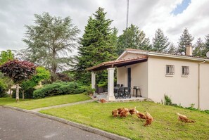 Ein eingeschossiges Haus mit überdachter Veranda, einem kleinen Garten und Hühnern auf der Wiese.