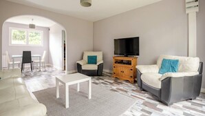 A living room with a sofa, armchair, TV stand, and a dining area with chairs and a table. - Peace 3 bed gîte (Chevanceaux)