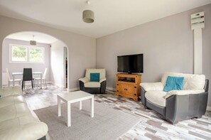 Ein Wohnzimmer mit einer Couch, Sesseln, einer TV-Konsole und einem Essbereich mit Stühlen und einem Tisch.