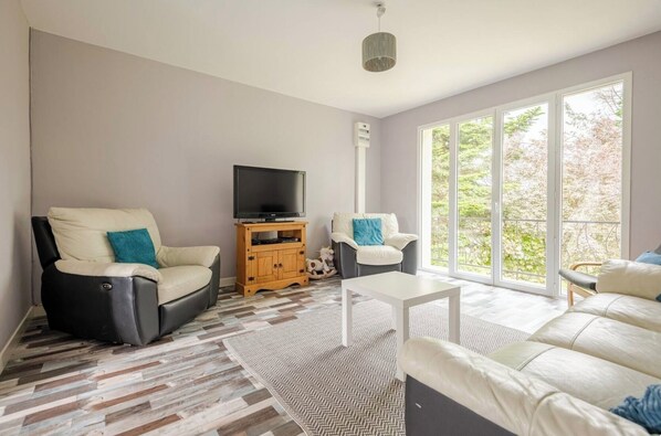 Ein Wohnzimmer mit Sofa, Sessel, TV-Schrank und Blick durch große Fenster.