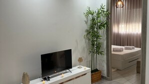 Ein modernes Wohnzimmer mit einem Flachbildfernseher auf einer weißen Konsole, einer großen Pflanze und einem Bett mit Baldachin.