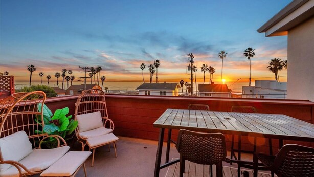 Une terrasse meublée avec des meubles en osier, une table et des chaises, d’où l’on peut admirer un coucher de soleil sur des palmiers et des bâtiments.