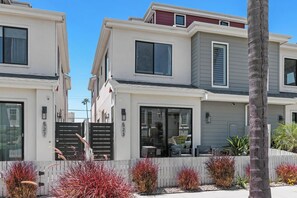 Un edificio residencial moderno de varios pisos con puertas numeradas, una entrada con portón y un jardín delantero ajardinado con diversos arbustos.