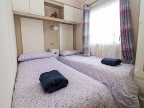 Quarto com duas camas, uma mesinha de cabeceira, uma prateleira com objeto decorativo, luminária na parede e janela com cortinas.