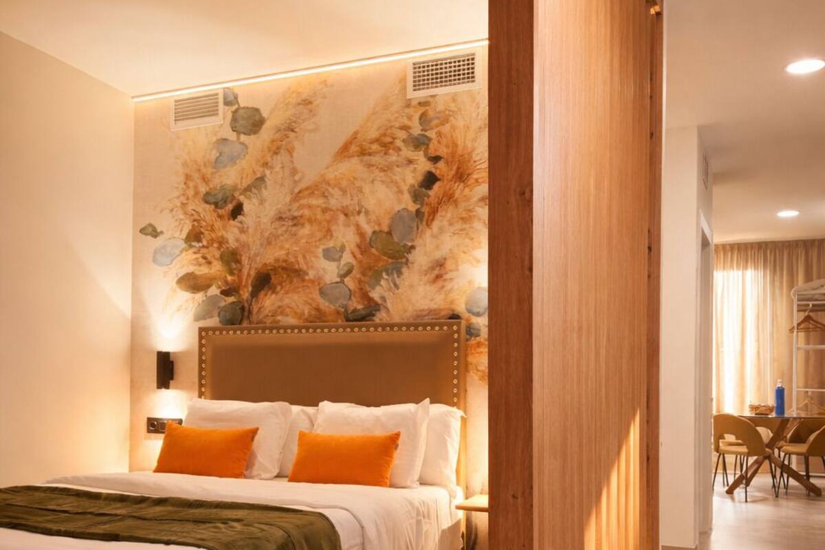 A modern hotel room with a wooden bed frame, a headboard with a decorative panel, and a dining area in the background.