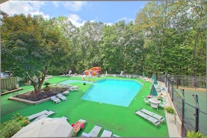 Ein rechteckiger Swimmingpool mit einer Rutsche, umgeben von grünem Kunstrasen und beschattet durch Sonnenschirme.