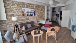 A living room with a stone wall, a sofa, a coffee table, a wicker chair, and a kitchen in the background. - Old fisherman's cottage completely renovated (Étel)