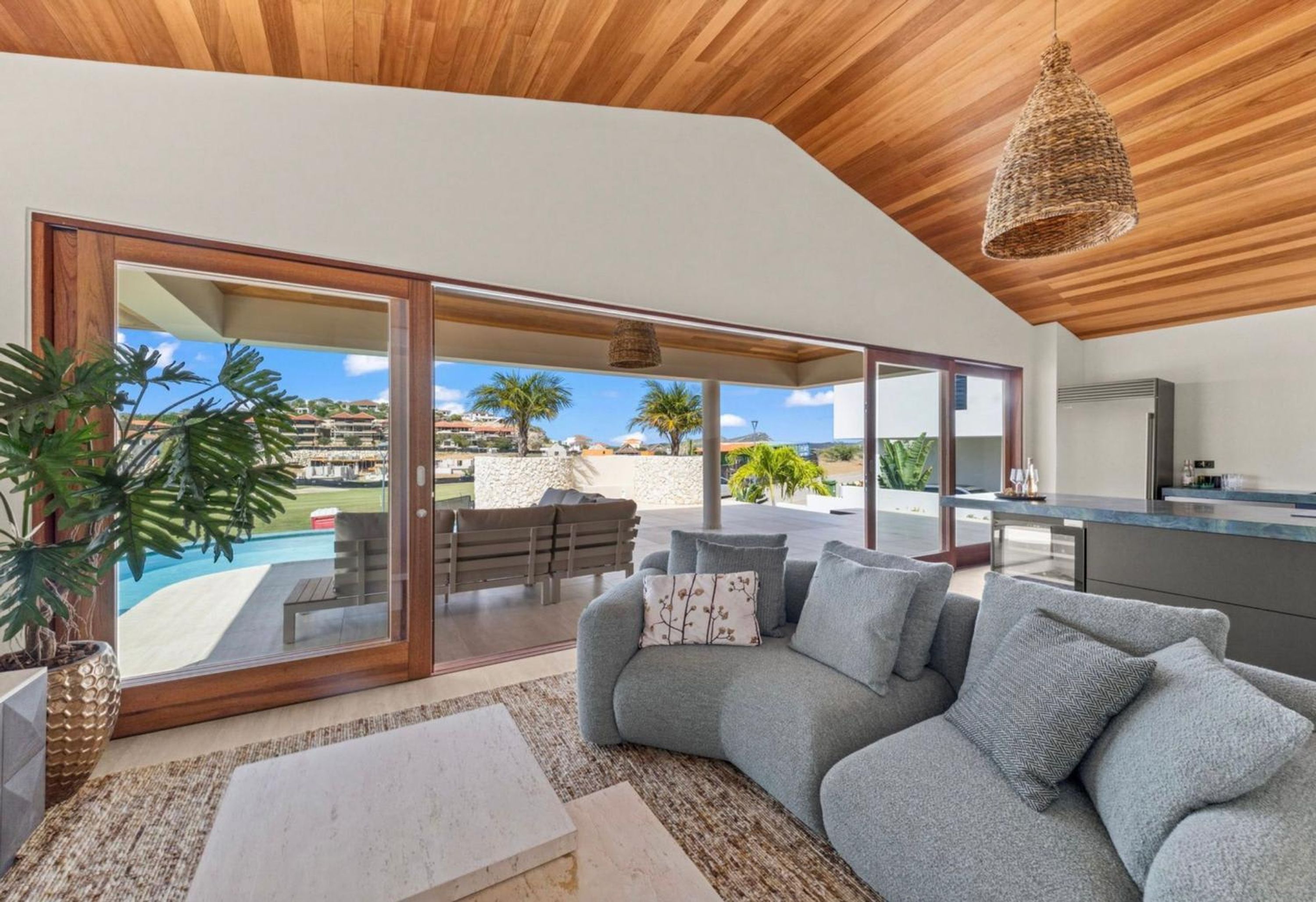 A modern living room with a grey sofa, a large window, and a view of a pool and palm trees.