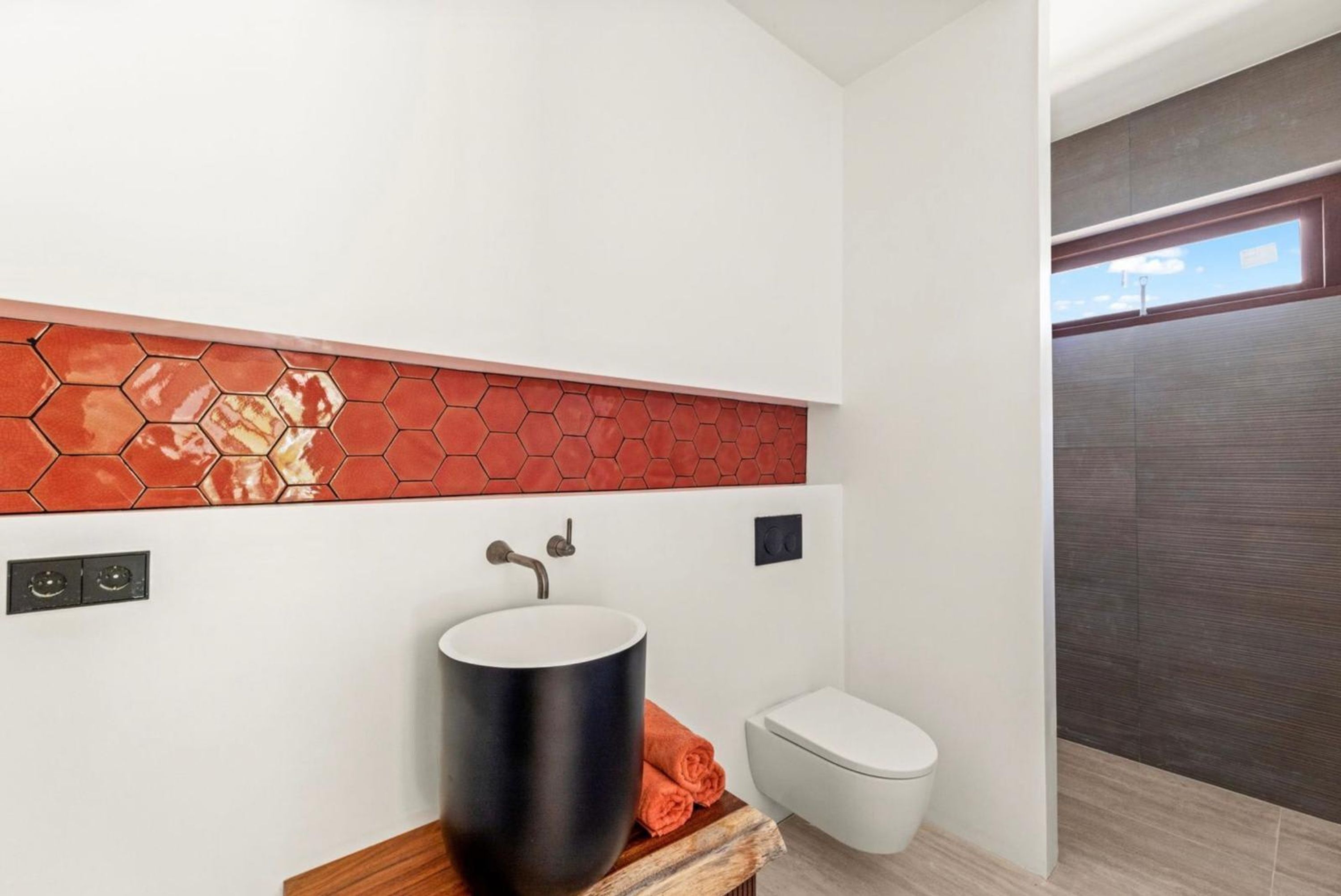 A modern bathroom with a hexagonal tile backsplash, a black vessel sink, a toilet, and a wooden vanity.