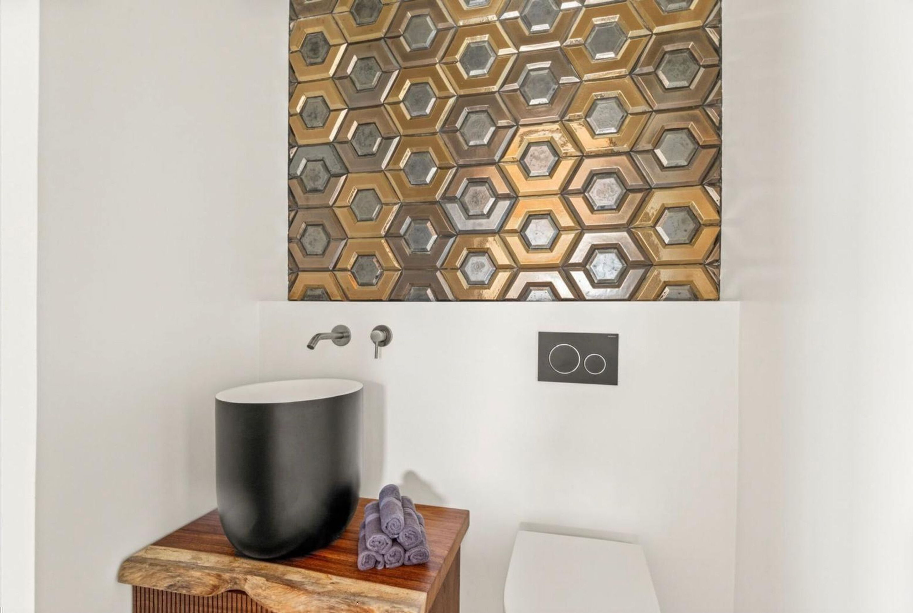 Ett badrum med en stor, svart tvättställ, ett sidobord i trä, hoprullade handdukar och en vägg med ett hexagonalt mönster.