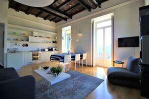 Ein modernes Wohnzimmer mit einer Couch, einem Couchtisch mit Pflanze, einem Essbereich mit Tisch und Stühlen, einer Küche mit weißen Schränken und Geräten sowie einem Balkon mit Blick.