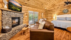Ein gemütliches Blockhauss-Interieur mit Kamin aus Stein, einem darüber montierten Fernseher, einem bequemen Sofa, einem Bett mit weißem Kopfteil und einem Deckenventilator.