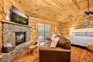 A cozy cabin interior with a stone fireplace, a television mounted above it, a comfortable brown sofa, a bed with a white headboard, and a ceiling fan. - Riverfront | Dog Friendly | Close to GSMNP (Townsend)