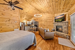 A log cabin interior with a bed, sofa, fireplace, kitchen area, and a ceiling fan. - Riverfront | Dog Friendly | Close to GSMNP (Townsend)
