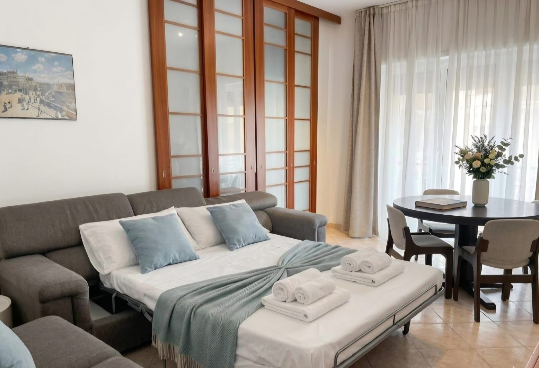 A modern bedroom with a sofa bed, a dining table with chairs, and a large window with curtains.