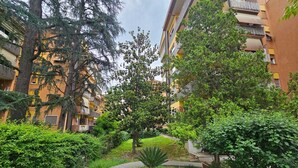 A pathway leading through a lush garden area with dense foliage and trees, surrounded by multi-story residential buildings. - Cozy apartment in Talenti - Well connected and quiet" (Roma)