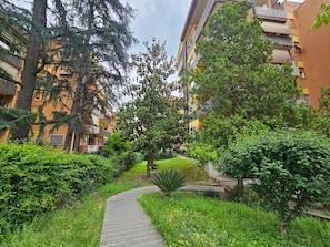 A pathway leading through a lush garden area with dense foliage and trees, surrounded by multi-story residential buildings. - Cozy apartment in Talenti - Well connected and quiet" (Roma)