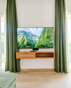 Ein Flachbildfernseher an der Wand montiert, unterhalb eine Holzablage, seitlich umrahmt von grünen Vorhängen, in einem Raum mit Holzboden.