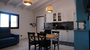 Una cuina moderna amb armaris blancs, una taula de dinar amb cadires, un sofà blau i dues làmpades penjants.