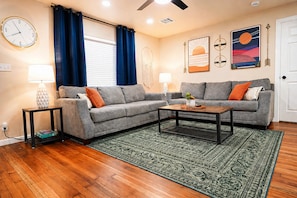 A living room with a grey sofa, a coffee table, a patterned rug, a clock, and wall art. - The Pinewood House | Near USAO + Close to Downtown (Chickasha)