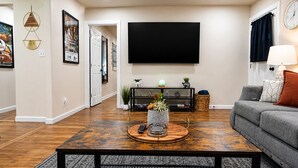 Una sala de estar con una televisión de pantalla plana, un sofá gris, una mesa de centro con objetos decorativos y una estantería.