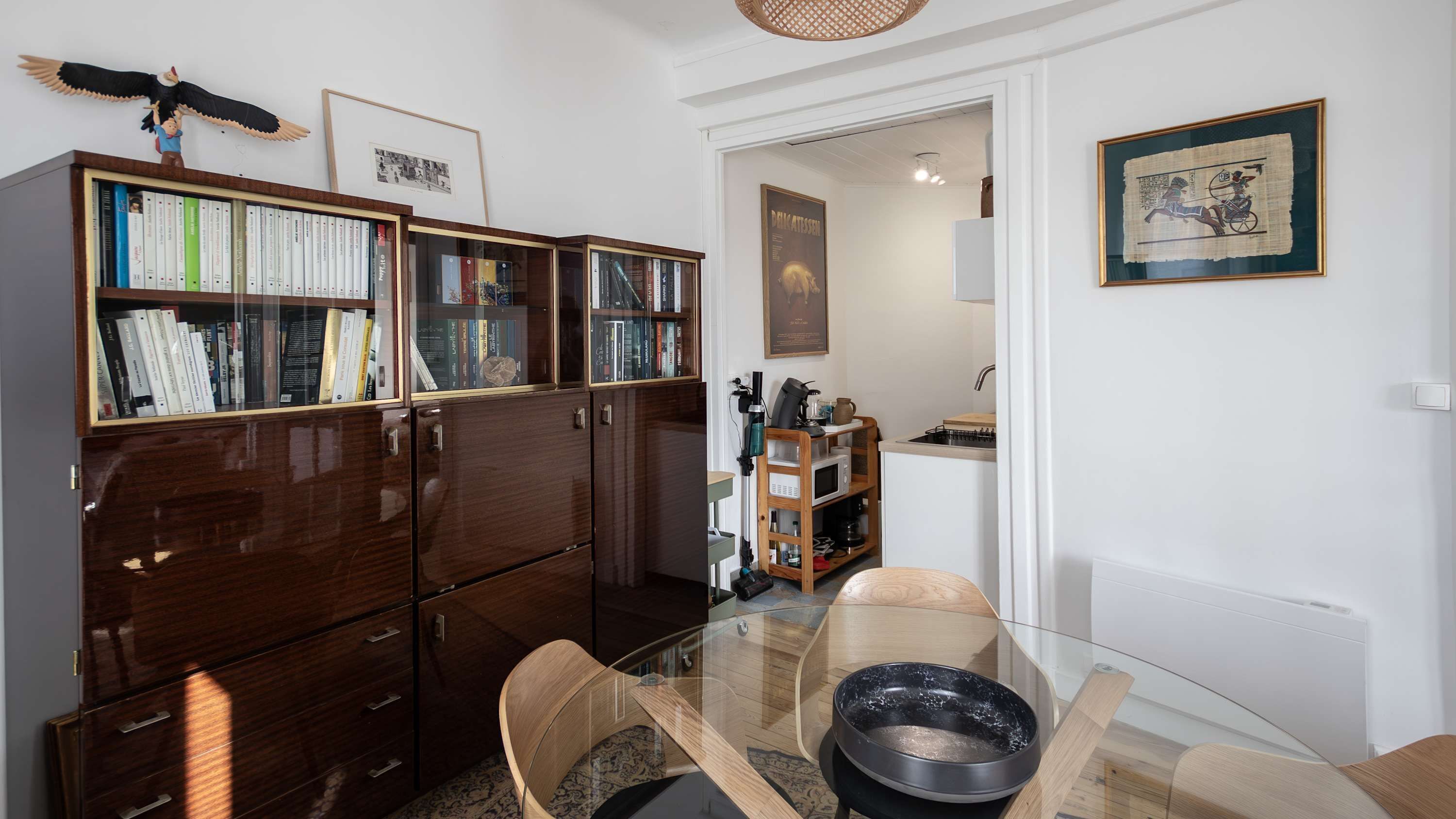 A room with a wooden bookshelf, a glass dining table, and a kitchen area visible through an open door.