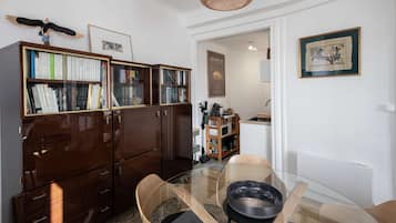 A room with a wooden bookshelf, a glass dining table, and a kitchen area visible through an open door.
