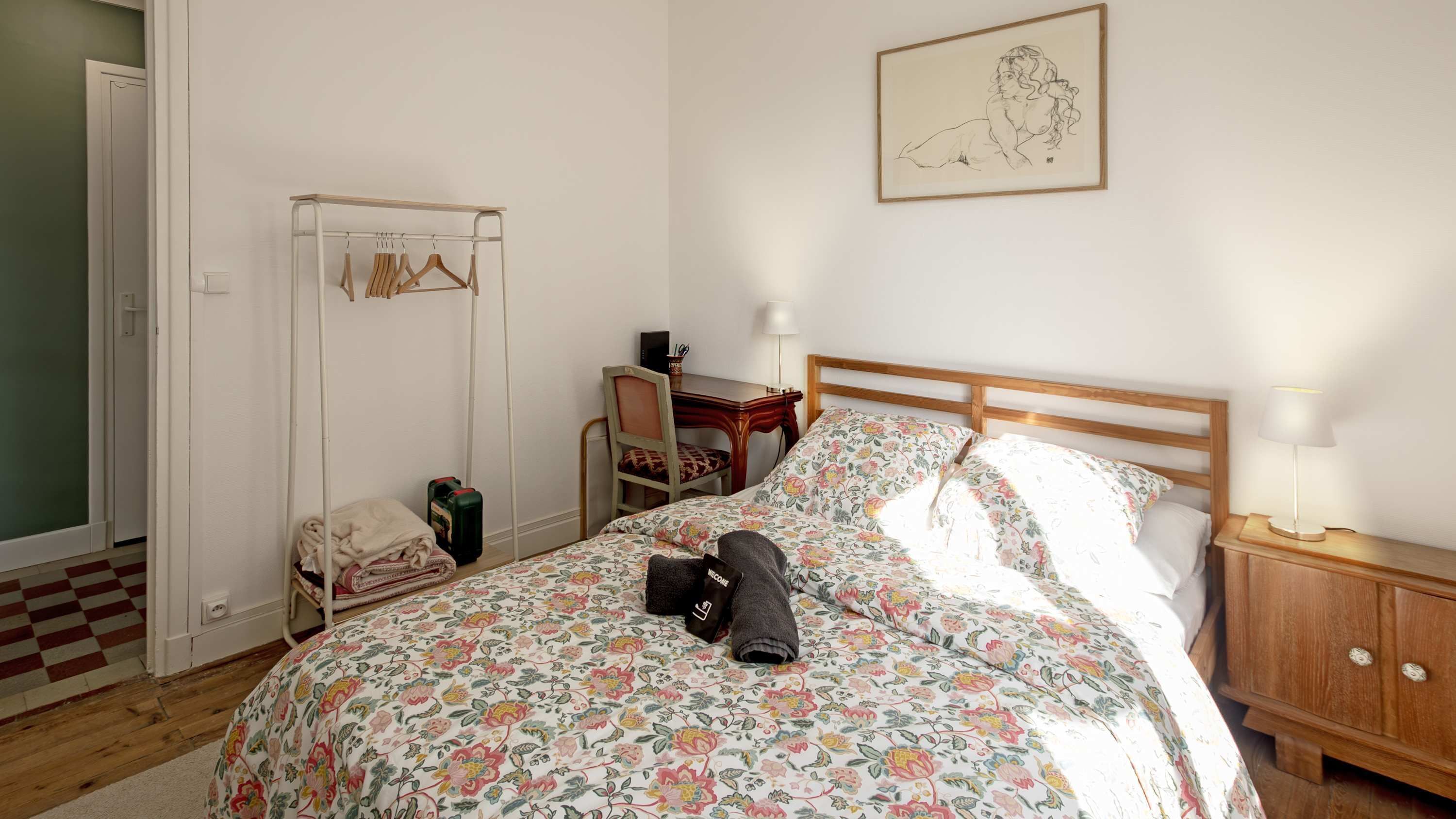 A bedroom with a bed, a desk, a chair, a coat rack, and a painting on the wall.