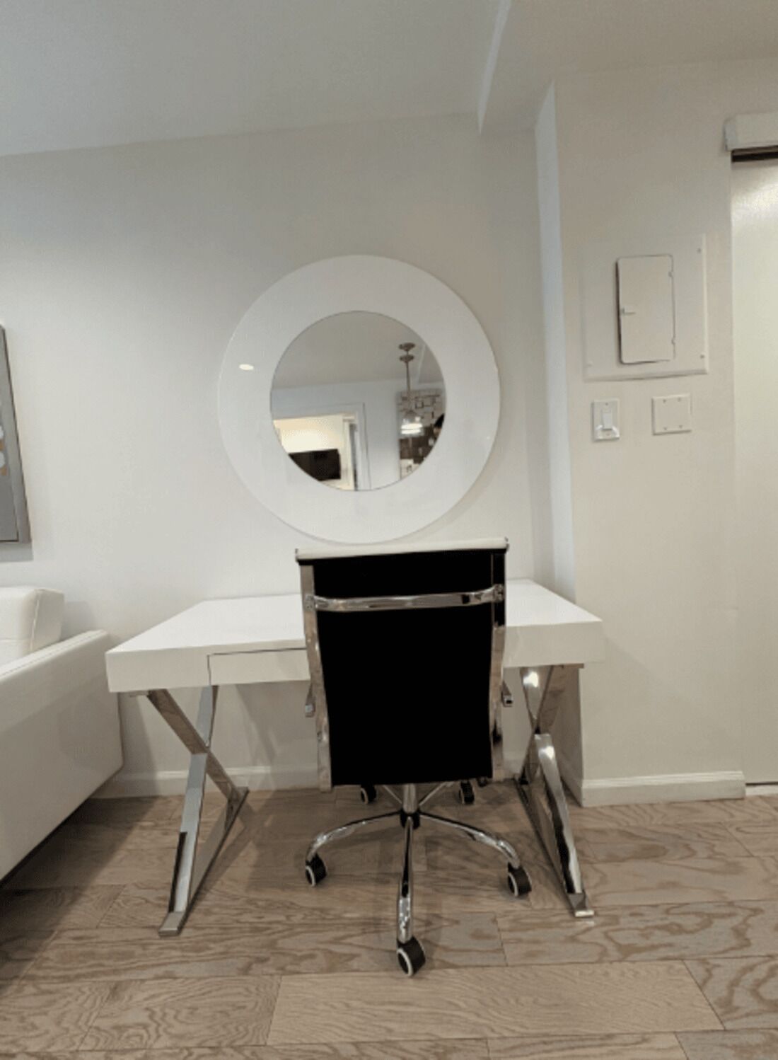 A white desk with a black chair, a round mirror on the wall, and a light switch.