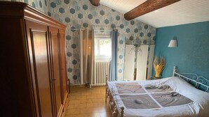 A bedroom with a wooden wardrobe, a bed with a patterned bedspread, a blue accent wall, and a sloped ceiling with exposed beams.