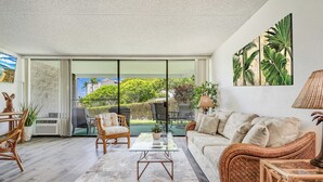 Amplia sala de estar con un gran ventanal corredero que da a un balcón, mobiliario de mimbre y decoración de temática tropical.